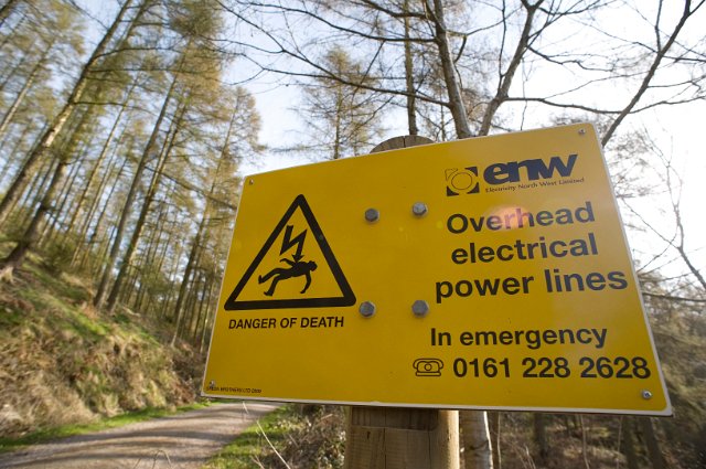 Yellow warning sigh for high voltage electricity cables overhead outdoors in woodland with an emergency contact number in case of a problem